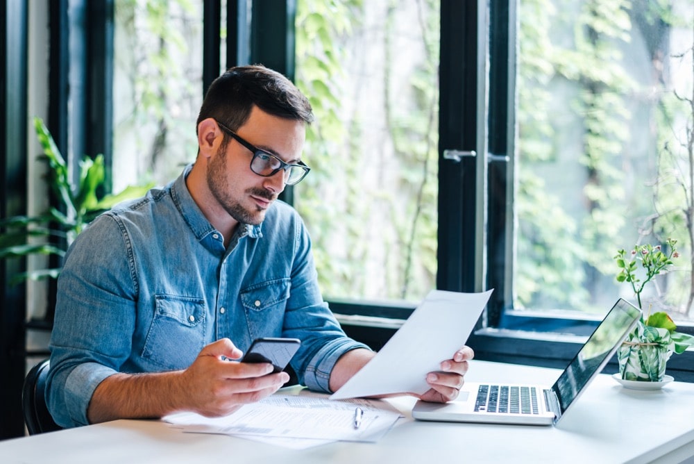 Serious pensive thoughtful focused young casual entrepreneur small business owner accountant bookkeeper in office looking at and working with laptop and income tax return papers and documents