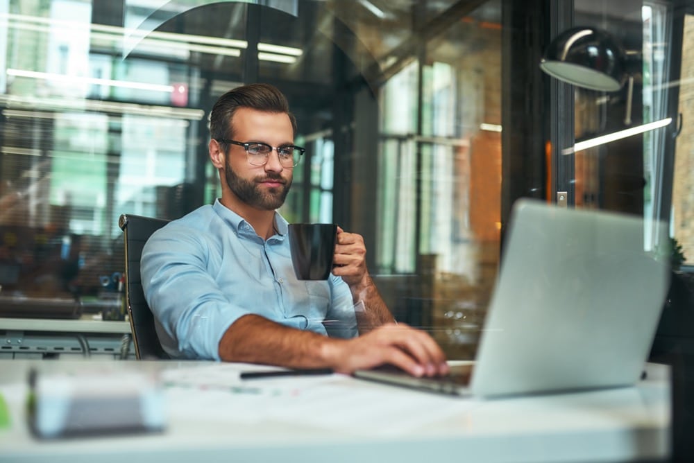 Morning coffee. Portrait of young and successful bearded man in eyeglasses holding cup of coffee and working with laptop while sitting at his working place